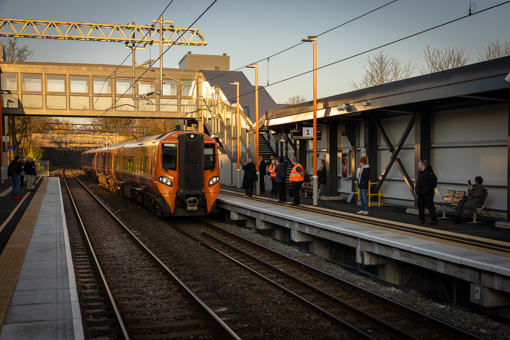 Train coming into station - a footbridge is over the track and passengers wait on the platform.